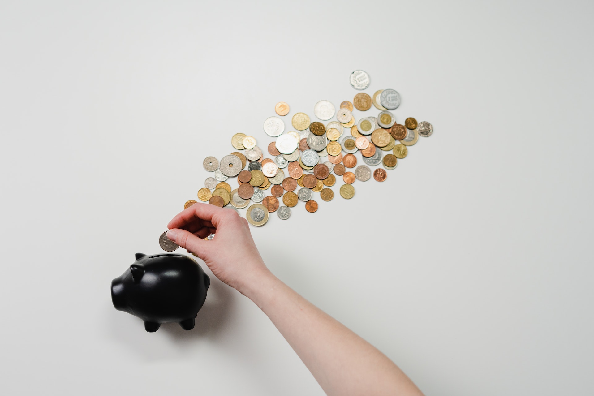 A top-down view of a person's hand as they place more money into a black piggy bank with many coins just on the outside of it.